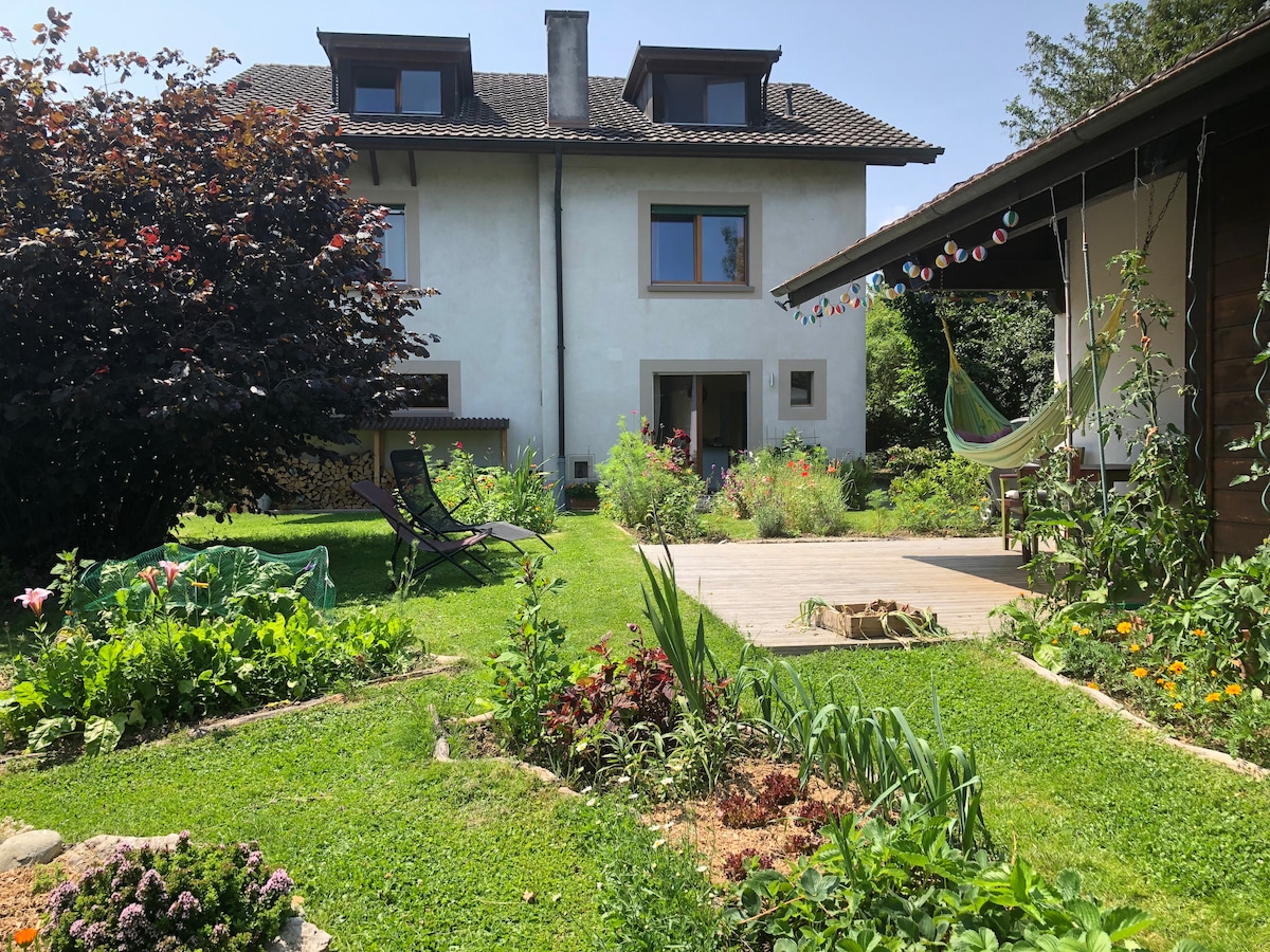 A serene garden area features lush greenery and colorful flowers surrounding a two-story building. A wooden deck extends into the yard, with seating arrangements visible. A hammock hangs nearby, and string lights add an inviting touch to the outdoor space.
