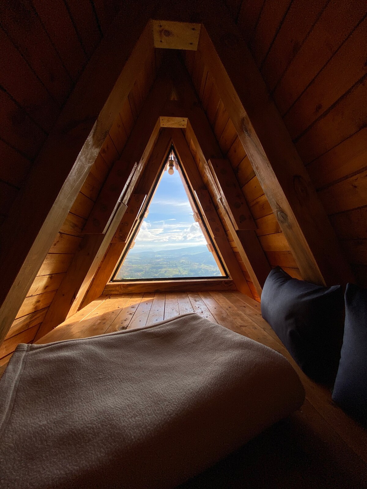 A triangular window framed by wooden walls offers expansive views of the landscape. A soft blanket is draped over a wooden surface, complemented by a cushioned area nearby. Natural light floods the space, highlighting the warm tones of the wood.