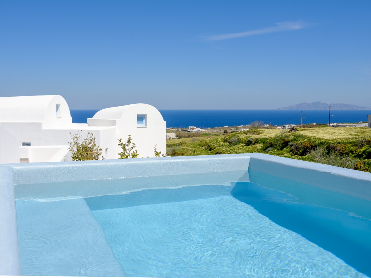 A private heated jacuzzi is positioned against a backdrop of the deep blue Aegean Sea. The clear water reflects the bright sky, enhancing the serene atmosphere. White Cycladic architecture is visible in the distance, surrounded by lush greenery.