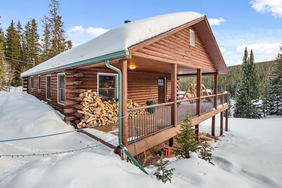 A log cabin is surrounded by snow-dusted terrain, with a large deck that offers views of the winter landscape. Stacked firewood is visible beside the cabin, and the roof is blanketed in snow, while tall evergreen trees frame the scene.
