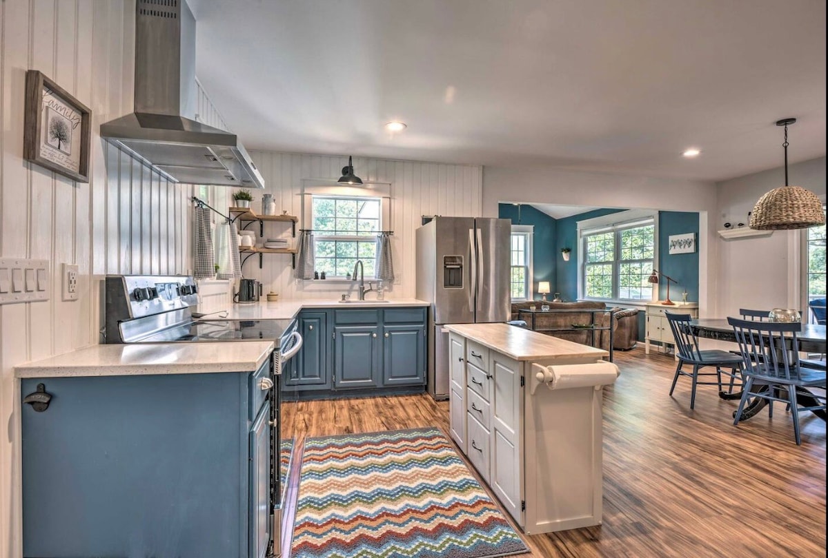 A spacious kitchen is presented, featuring stainless steel appliances and a central island with seating. Light countertops contrast with blue cabinetry, and a colorful woven rug provides warmth underfoot. Natural light filters through multiple windows, illuminating the vibrant dining area visible in the background.