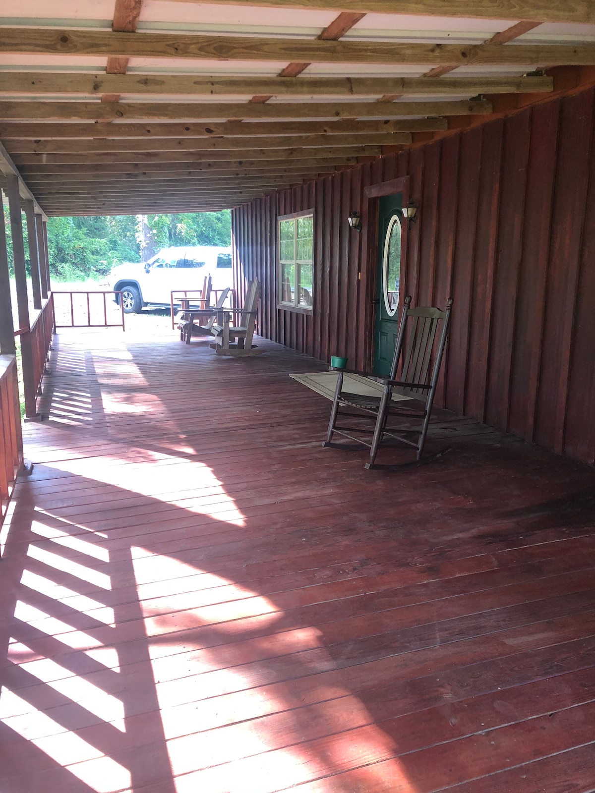 A covered porch features wooden flooring and a rustic ambiance. Two rocking chairs are positioned on the left, with light and shadow patterns created by the overhead beams. A green door is situated in the center, accompanied by large windows that allow natural light to enter.