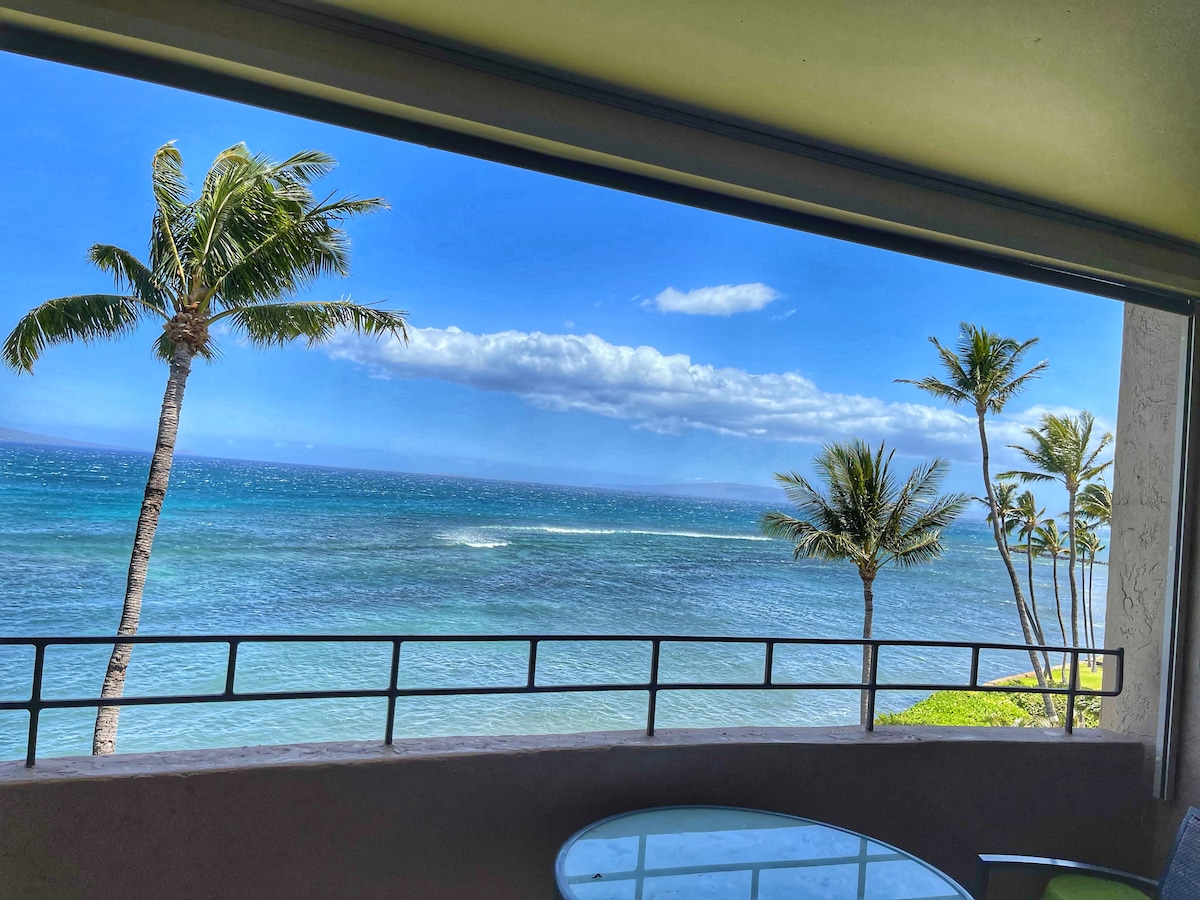 A scenic ocean view is framed by large windows, showcasing palm trees swaying gently in the breeze. The tranquil waters of Ma'alaea Bay reflect the clear blue sky, inviting relaxation from a private lanai with a circular table set for outdoor enjoyment.