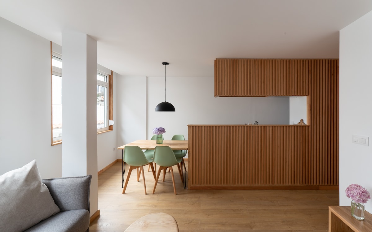 A modern living space features a light wooden floor and a stylish dining area. A round table is surrounded by green chairs, while a wooden partition separates the kitchen. Large windows allow natural light to fill the room, complementing the neutral color palette.