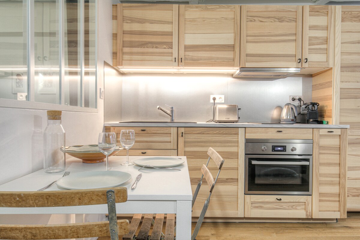 A functional kitchen area features light wood cabinetry and modern appliances. A small dining table is set for two, accompanied by simple wooden chairs. Natural light brightens the space, reflecting off the stainless steel surfaces and creating a warm and inviting environment.