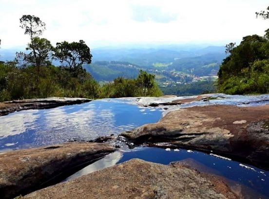 A panoramic view of a natural landscape is presented, featuring a rocky outcrop with a calm water pool reflecting the sky. Lush greenery surrounds the area, and rolling hills stretch out in the distance, showcasing a mix of mountainous terrain and a small town nestled below.