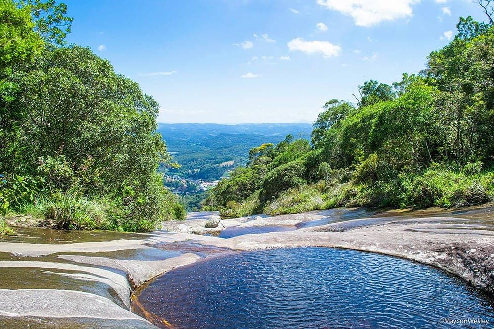 A serene view of a natural landscape shows gentle slopes and rocky formations leading to a large, tranquil pool. Green vegetation surrounds the area, providing a lush backdrop against a clear blue sky. The distant hills can be seen in the horizon.