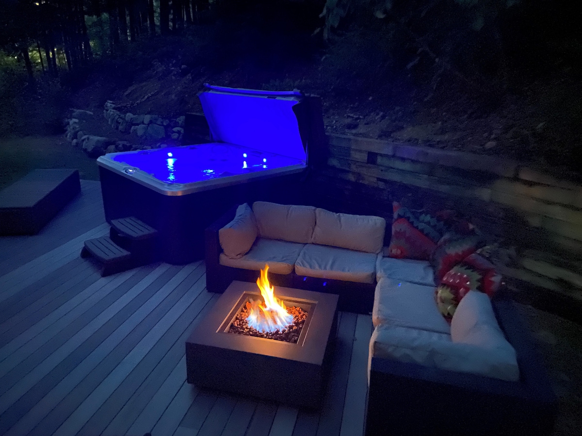An outdoor space is presented with a large hot tub emitting a soft blue glow, alongside a fire pit surrounded by a sectional sofa. Cozy cushions enhance comfort, while the surrounding night adds a serene ambiance to the deck.