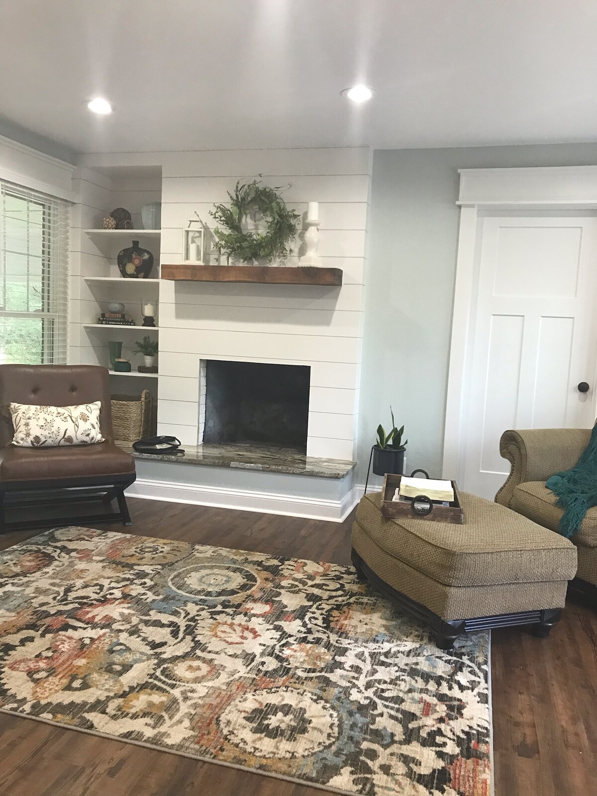 A cozy living space is presented with a plush beige chair and a black rocking chair. A decorative area rug in warm tones covers the floor. A white shiplap wall features a fireplace and a wooden shelf adorned with decorative items, while natural light filters through the nearby window.