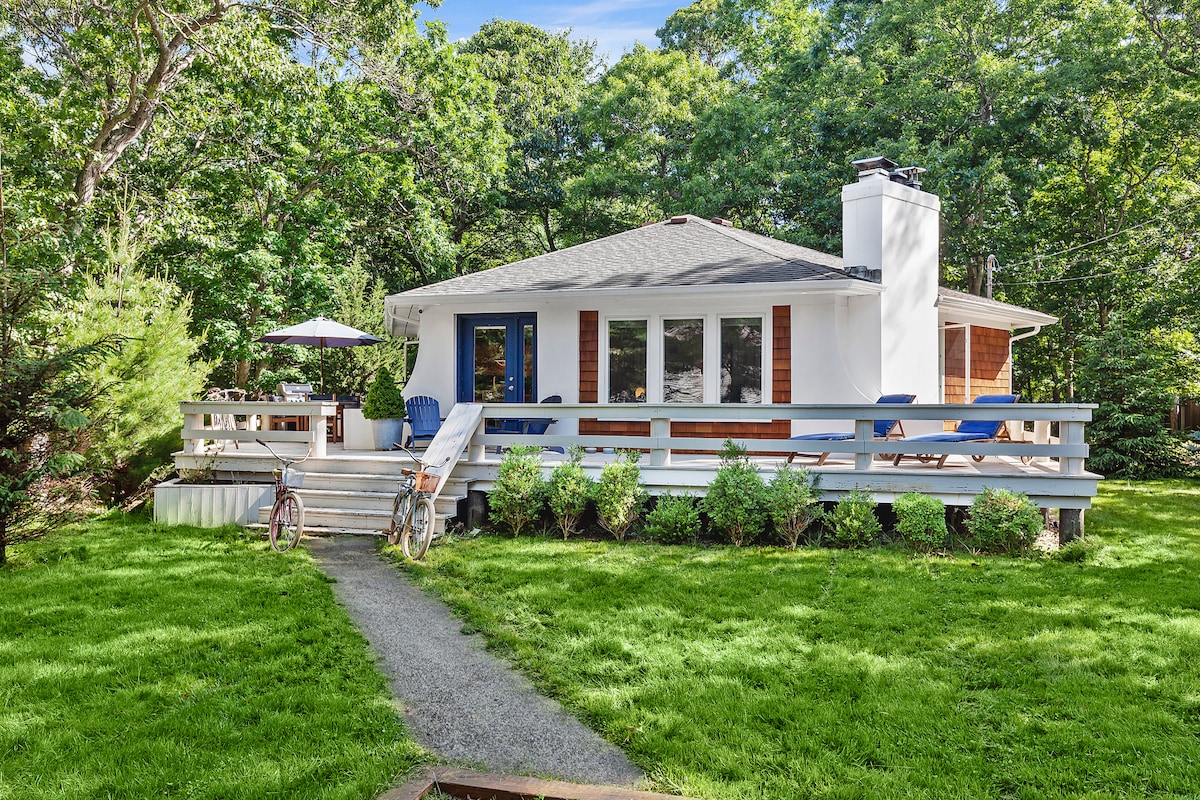 A hexagonal-shaped bungalow with white exterior and large front windows, sits amidst lush greenery. A pathway lined with grass leads to the entrance. Two blue chairs and an umbrella can be seen on the deck, providing a space to relax outdoors.