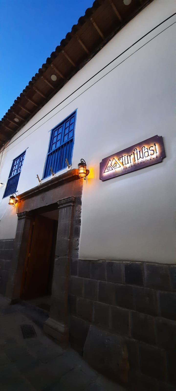 The exterior of a charming building is captured in twilight, featuring a welcoming entrance framed by stone. Blue wooden windows contrast against the white wall, while decorative lighting adds subtle illumination. A sign bearing the establishment's name is prominently displayed, enhancing the inviting appearance.