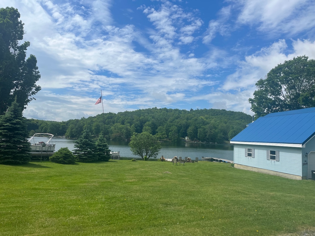 A serene view of the lake is framed by lush greenery and a spacious lawn. A charming blue cottage is positioned to the right, with a bright blue roof. An American flag waves gently in the distance, while a small dock is visible along the water's edge.