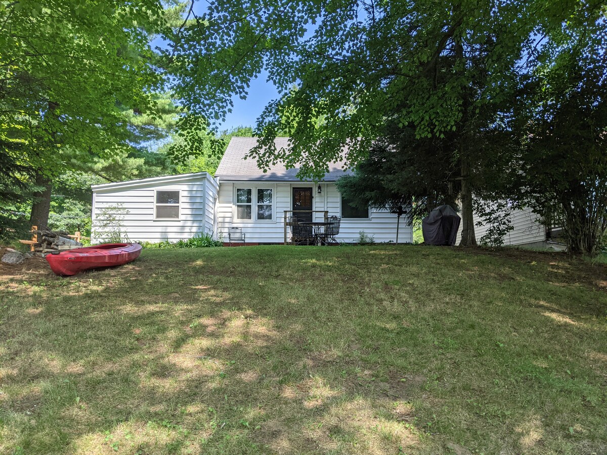 The exterior view of the house reveals a charming single-story structure surrounded by lush green trees. A spacious yard is visible, featuring a red kayak on the grass and a patio table with chairs near the entrance, offering a welcoming outdoor space.