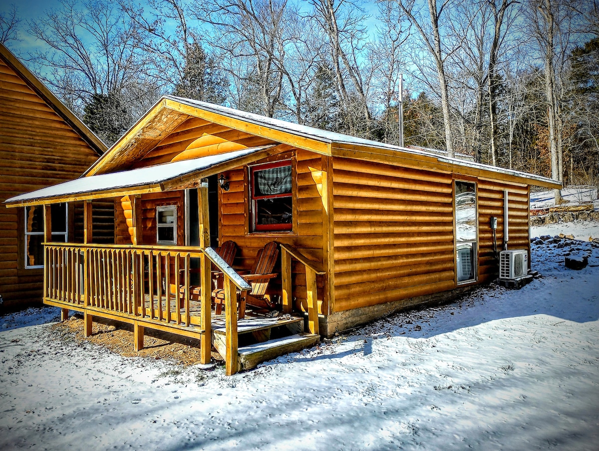 A rustic cabin features log siding and a welcoming porch adorned with two wooden chairs. The surrounding area is blanketed in snow, with trees in the background suggesting a tranquil, natural setting. A modern air conditioning unit is mounted on the side of the cabin.