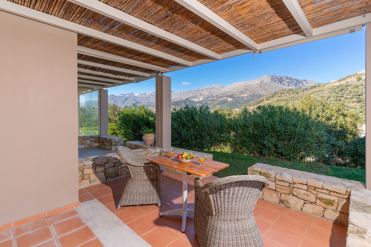 A shaded outdoor terrace offers a rustic wooden table surrounded by woven chairs. The view includes lush greenery and distant mountains under a clear blue sky, creating a serene setting for meals or relaxation.