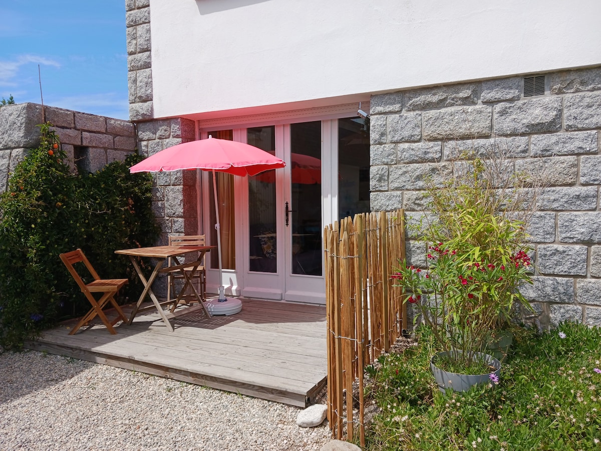 A wooden deck area features a small table and two chairs, accompanied by a bright pink umbrella. Surrounding greenery adds a natural touch, while a low bamboo fence creates privacy. The nearby stone wall complements the serene outdoor space.