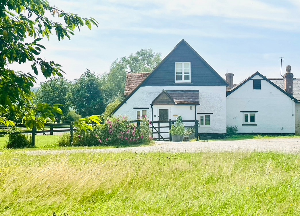A charming cottage, featuring a combination of traditional and modern architecture, stands amidst lush greenery. The exterior is highlighted by white walls and dark timber accents, with a welcoming front door and surrounding flower beds adding to its appeal.