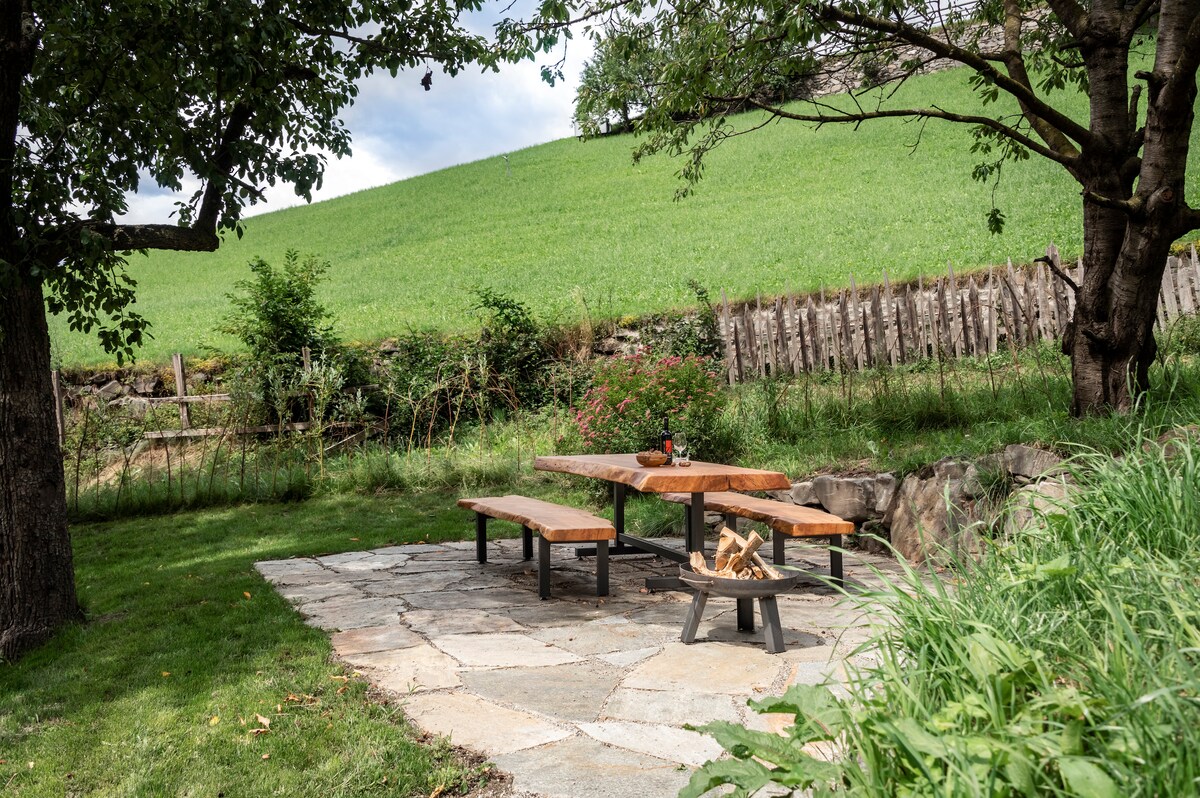 An outdoor seating area features a large wooden table with benches, positioned on a stone patio. A fire pit is located in the center. The area is surrounded by greenery and offers a view of a grassy hillside.