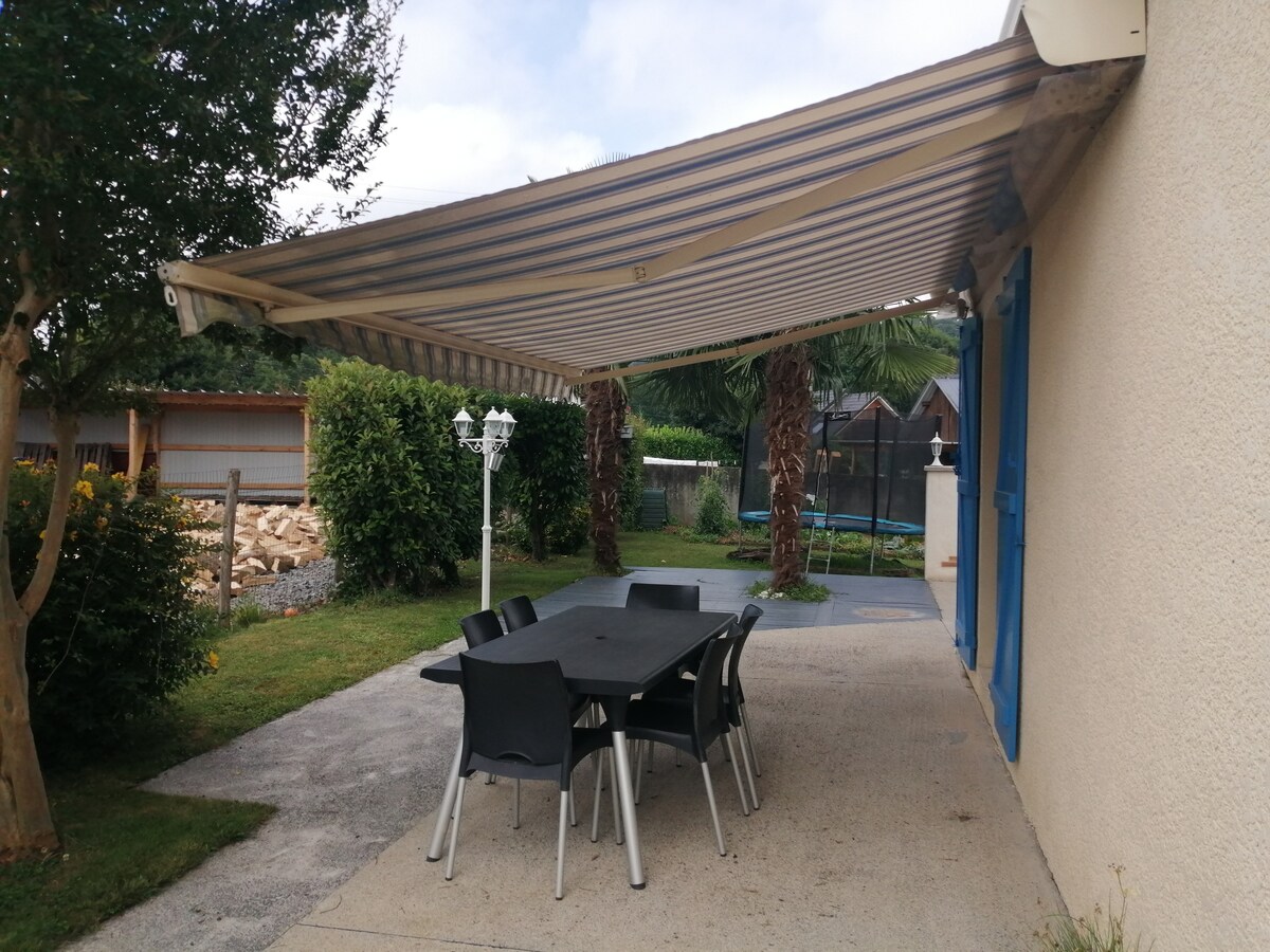 An outdoor dining area features a large table surrounded by six chairs, sheltered by a striped awning. Lush greenery and a trampoline are visible in the background, complemented by landscaped foliage and a stone wall, creating a serene environment.