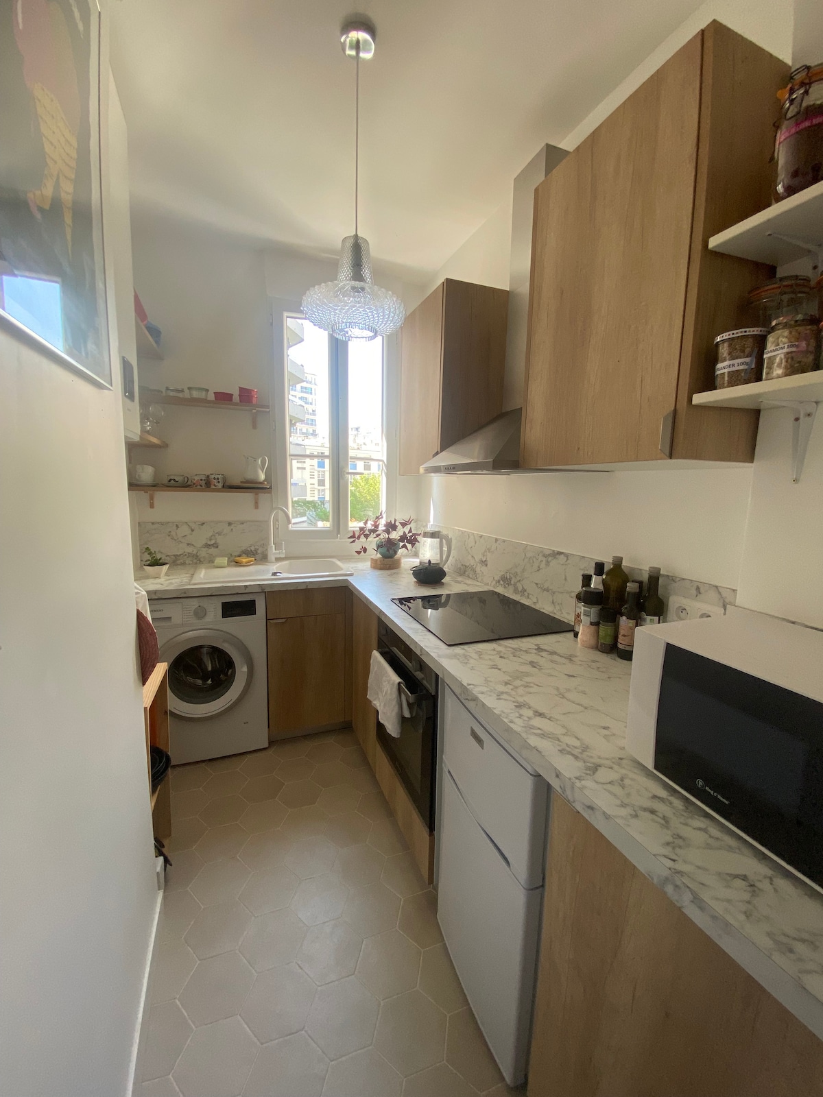 A well-equipped kitchen features modern wooden cabinetry and marble countertops. A washing machine is positioned beside the sink. Natural light enters through the window, illuminating the space. Open shelves display jars, and appliances include a microwave and a compact refrigerator.