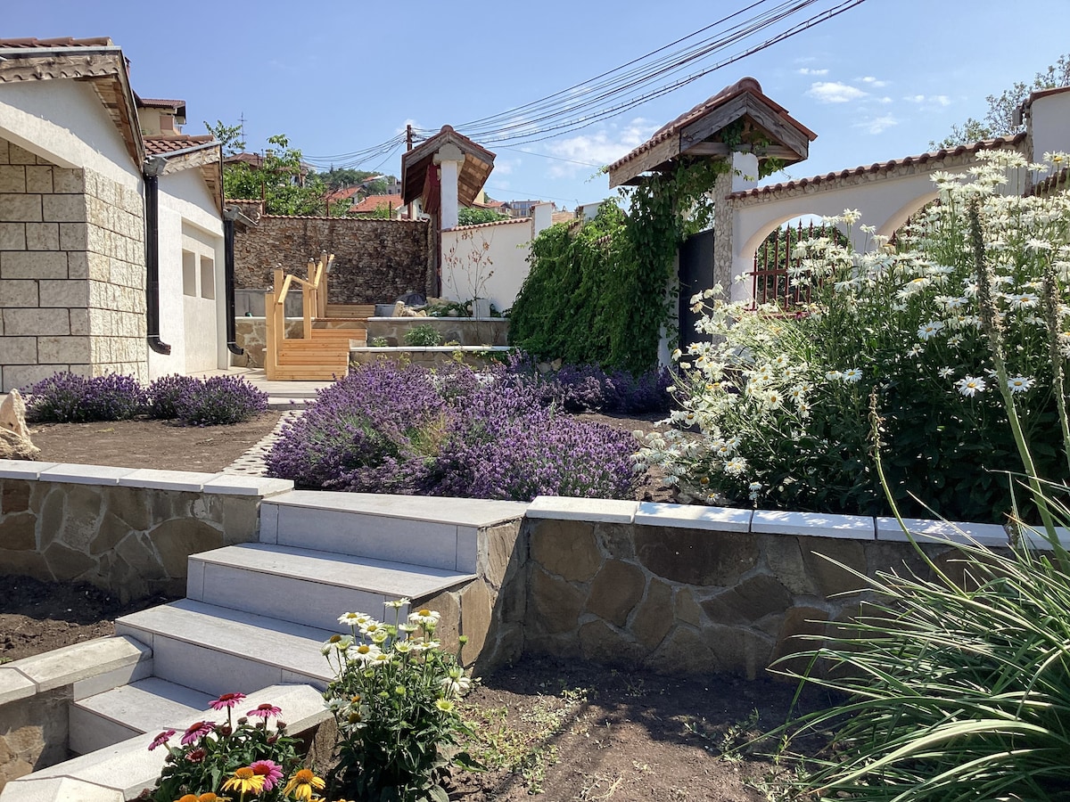 A landscaped outdoor area features a stone pathway leading to a wooden seating area amidst vibrant flower beds filled with lavender and other blooms. Lush greenery surrounds the space, with well-defined garden borders and rustic styled structures visible in the background.