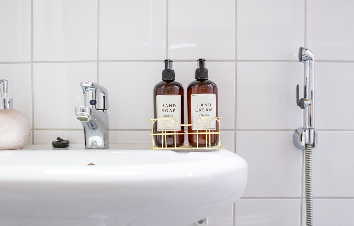 The bathroom sink area features a sleek white basin with a modern faucet. A gold wire caddy holds two amber bottles labeled 'Hand Soap' and 'Hand Cream,' adding a touch of elegance to the space. Clean, white tiles create a fresh backdrop.