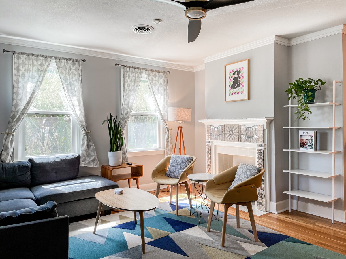 The living area features a cozy sectional sofa paired with stylish armchairs. Natural light enters through two large windows, illuminating the space. A modern coffee table is placed on a colorful area rug, accentuating the neutral tones of the walls and furnishings.