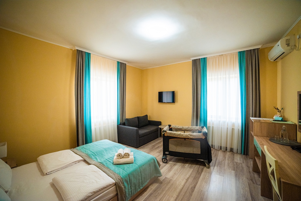 A well-lit room features a spacious bed with light-colored linens and neatly arranged towels. A comfortable sofa is positioned opposite a wall-mounted television. Large windows with sheer curtains allow natural light to brighten the room, while a stylish wooden console holds additional amenities.