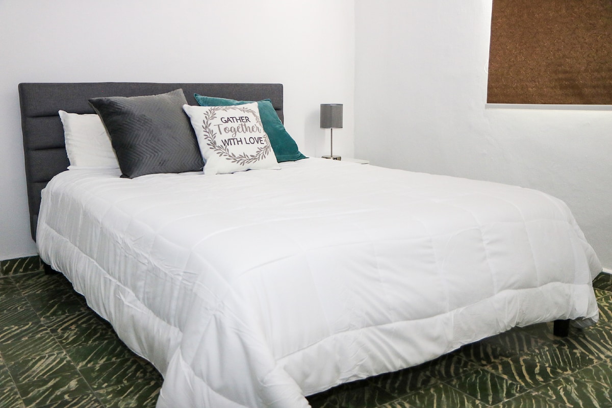 A spacious bed with a crisp white comforter is positioned against a grey upholstered headboard. Decorative pillows in various textures add subtle interest. A bedside lamp sits on a white nightstand, and natural light filters in softly through a window with a dark brown covering.