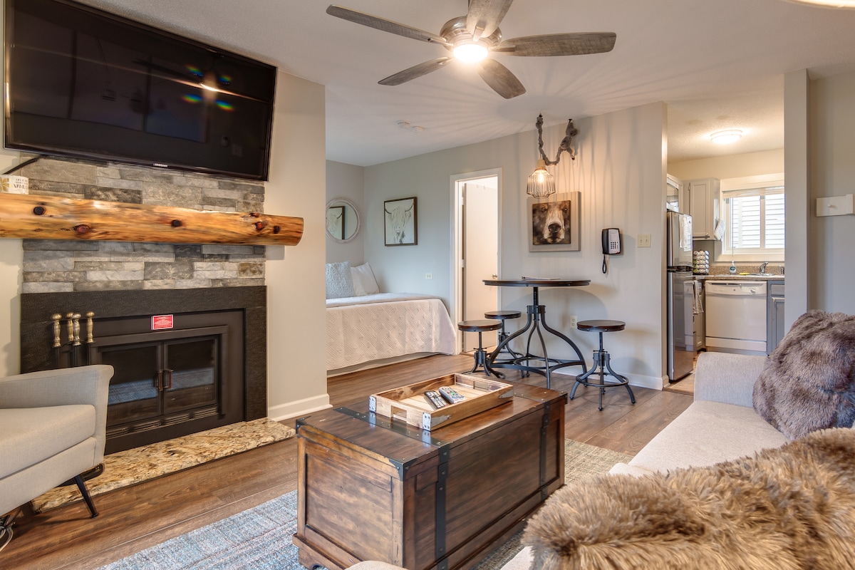 A warm living area is highlighted by a stone fireplace beneath a flat-screen TV. Light wood beams feature a rustic charm. A small dining table for two is present, alongside a cozy couch and a textured rug, creating an inviting space for relaxation.