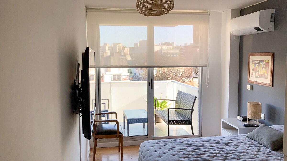 A spacious room is equipped with a comfortable bed and a flat-screen TV mounted on the wall. Natural light fills the space through large glass doors that lead to a private balcony, featuring outdoor seating and greenery.
