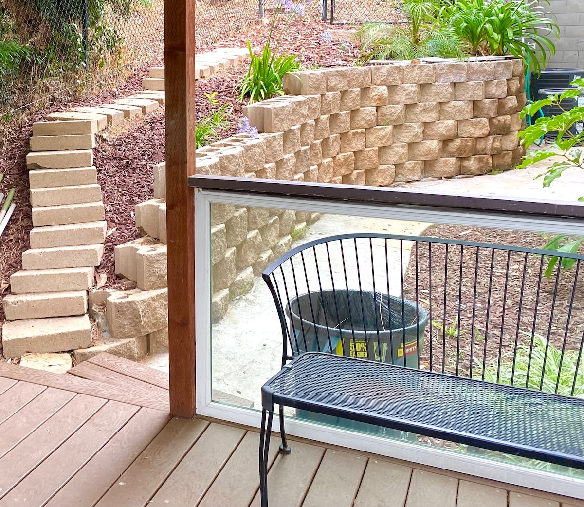 A private patio area features a metal bench overlooking a landscaped yard with terraced garden beds. A path, lined with steps, leads up the hill. Natural greenery surrounds the space, providing a tranquil atmosphere. A large container is visible for waste disposal.
