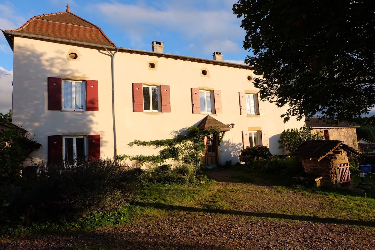 The exterior of a renovated historic house is shown, featuring light-colored walls and traditional wooden shutters. A covered entrance is framed by greenery, while a tranquil garden area with small structures and chairs can be seen in the foreground.