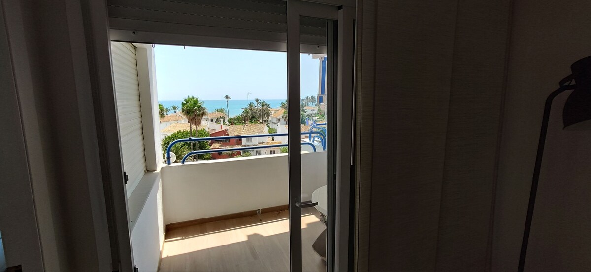A view from an open door showcases a balcony with railings and glimpses of the ocean. The scene includes palm trees and coastal buildings in the distance, providing a sense of connection to the surrounding environment.
