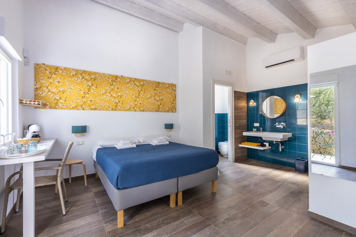 An elegant room with a spacious bed is highlighted by a vibrant yellow wall accent. A cozy sitting area is present along with a modern bathroom featuring a large mirror. Natural light enters through a window, revealing a view of a flowering courtyard.