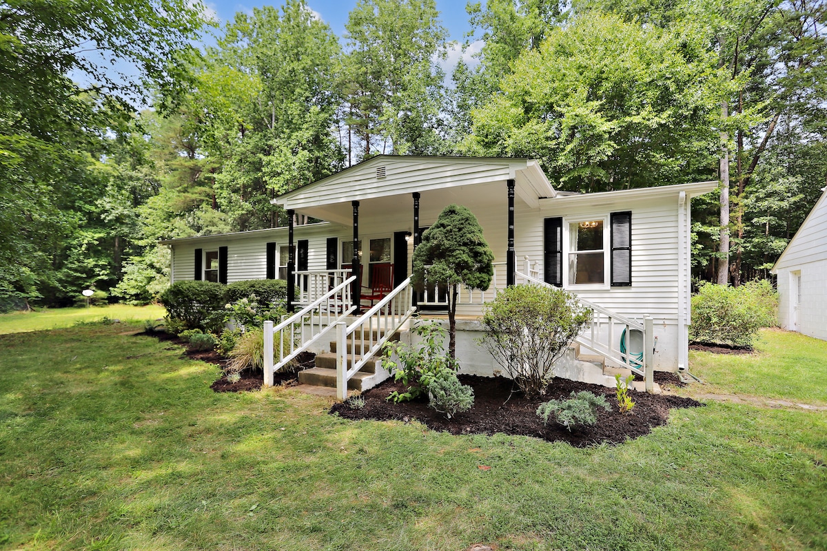 A newly renovated home is showcased with a welcoming front porch. Lush greenery surrounds the property, complemented by well-maintained shrubs and trees. The exterior features a light-colored siding with dark shutters and a manicured lawn leading up to the entrance.