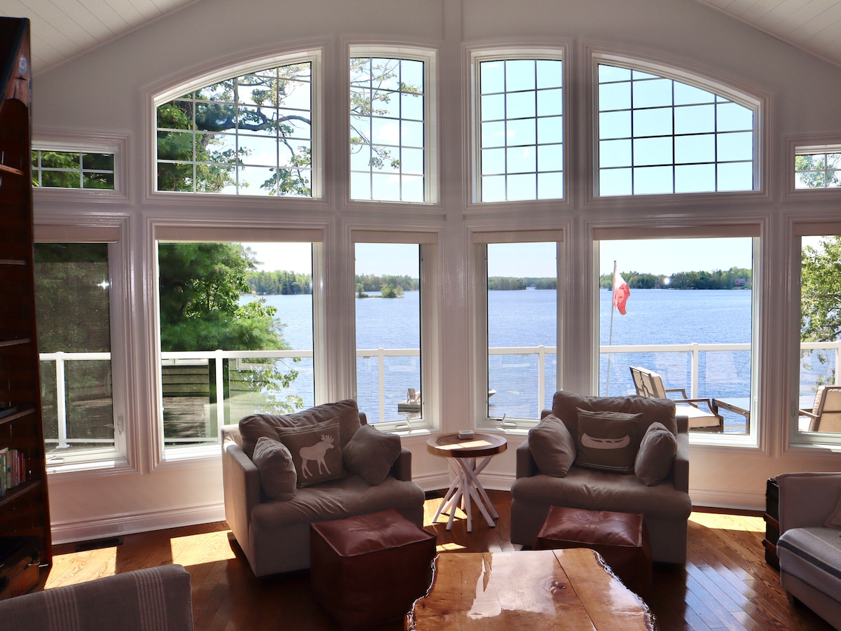 A spacious family room features floor-to-ceiling windows that provide a panoramic view of the lake. Comfortable seating includes a light-colored couch and two large club chairs, arranged around a wooden coffee table. Natural light enhances the airy atmosphere while a serene outdoor setting invites relaxation.