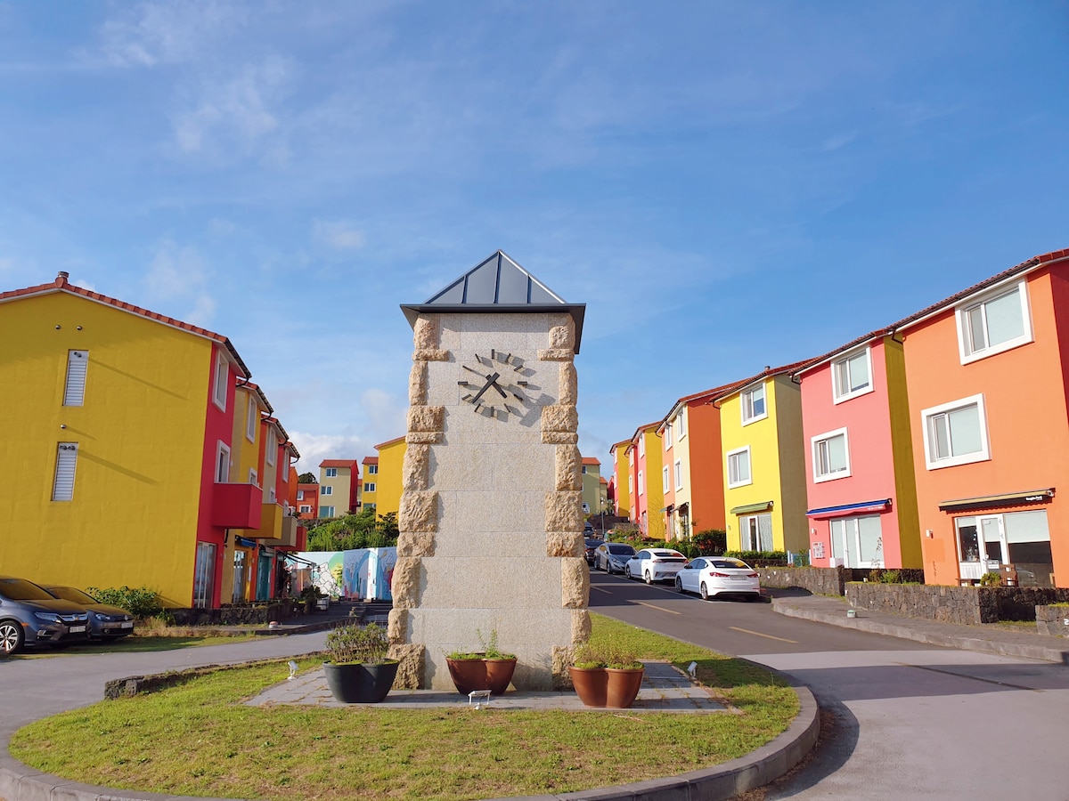 A stone clock tower stands prominently at the center of the roundabout, surrounded by brightly colored buildings in yellow and pink. Potted plants enhance the welcoming entrance, while neatly parked cars line the circular driveway, indicating a vibrant community atmosphere.