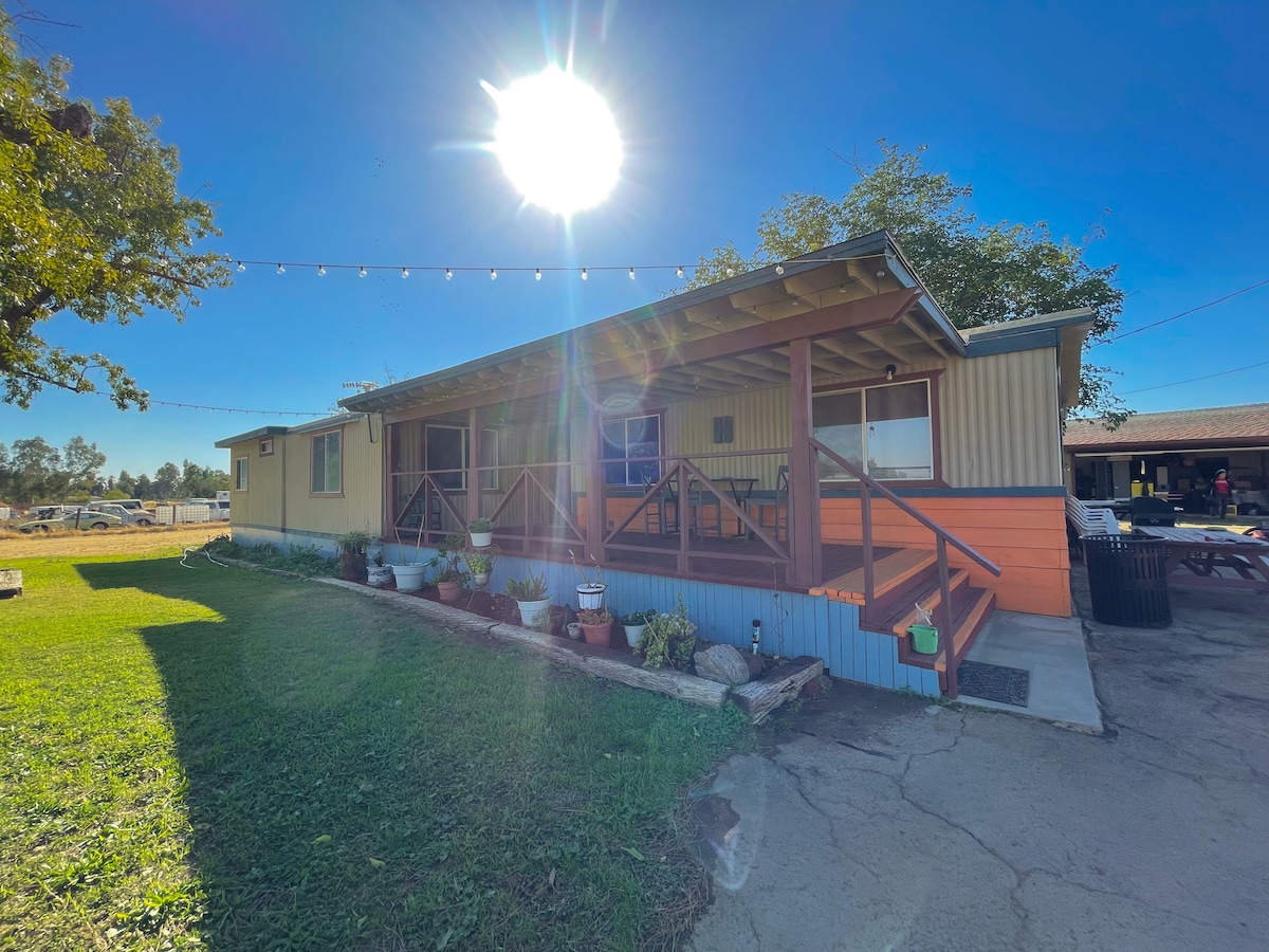 A single-story ranch home is shown under clear skies, featuring a wooden porch with railing and potted plants. Natural sunlight creates an inviting atmosphere, illuminating the exterior and surrounding grassy area.