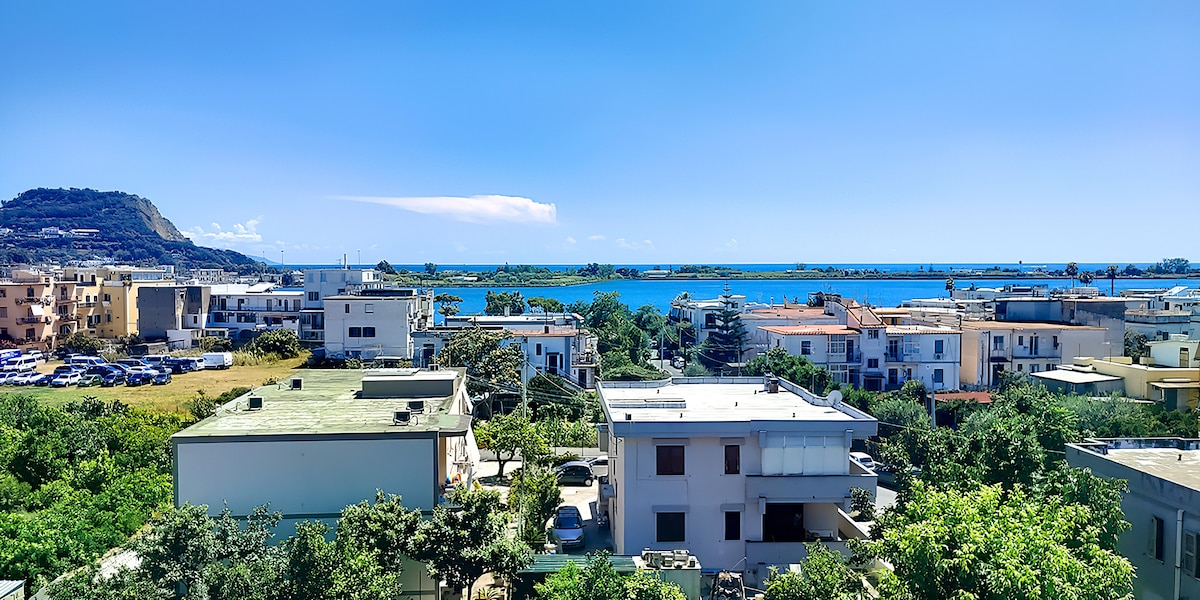 A panoramic view reveals a blend of residential buildings and greenery, with the serene blue water in the background. The coastline stretches towards the horizon, framed by distant hills under a clear blue sky.
