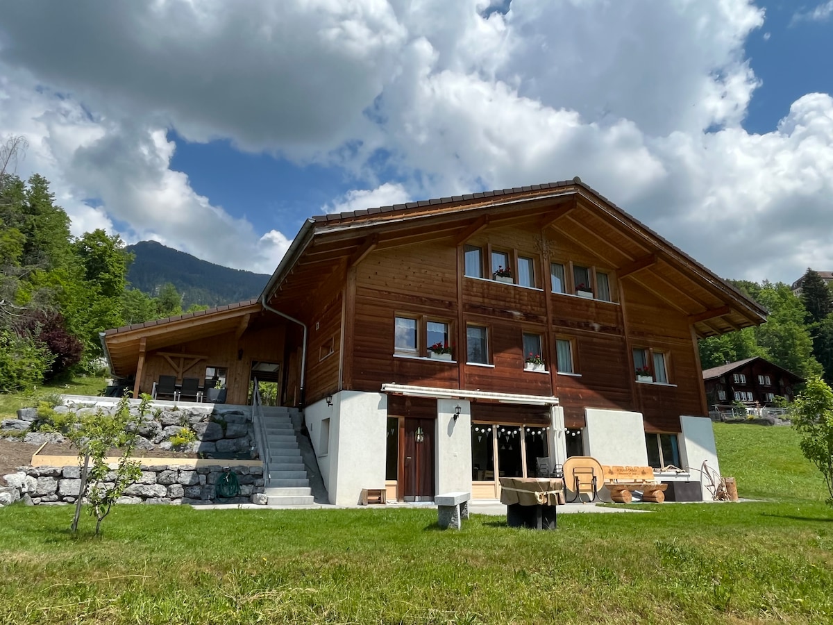 The chalet's exterior showcases a combination of wood and stone elements under a partly cloudy sky. The two-story building features large windows and a spacious entryway. A grassy area surrounds the structure, with outdoor seating visible and stairs leading to the entrance.