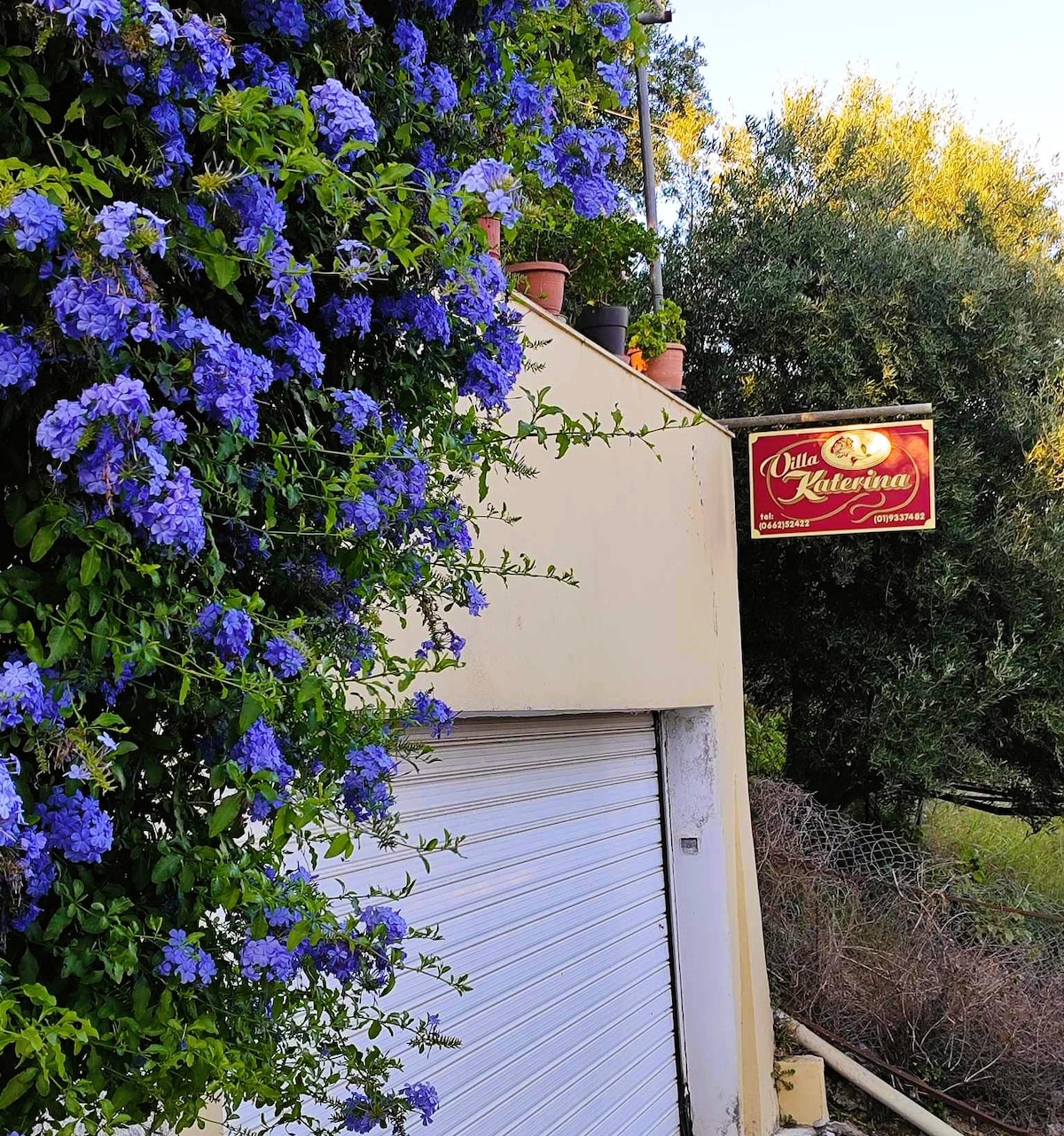 Purple flowering vines cascade along a cream-colored wall, leading to a sign that reads 'Villa Katerina.' A garage door, partially visible, complements the structure, surrounded by lush greenery typical of the area.