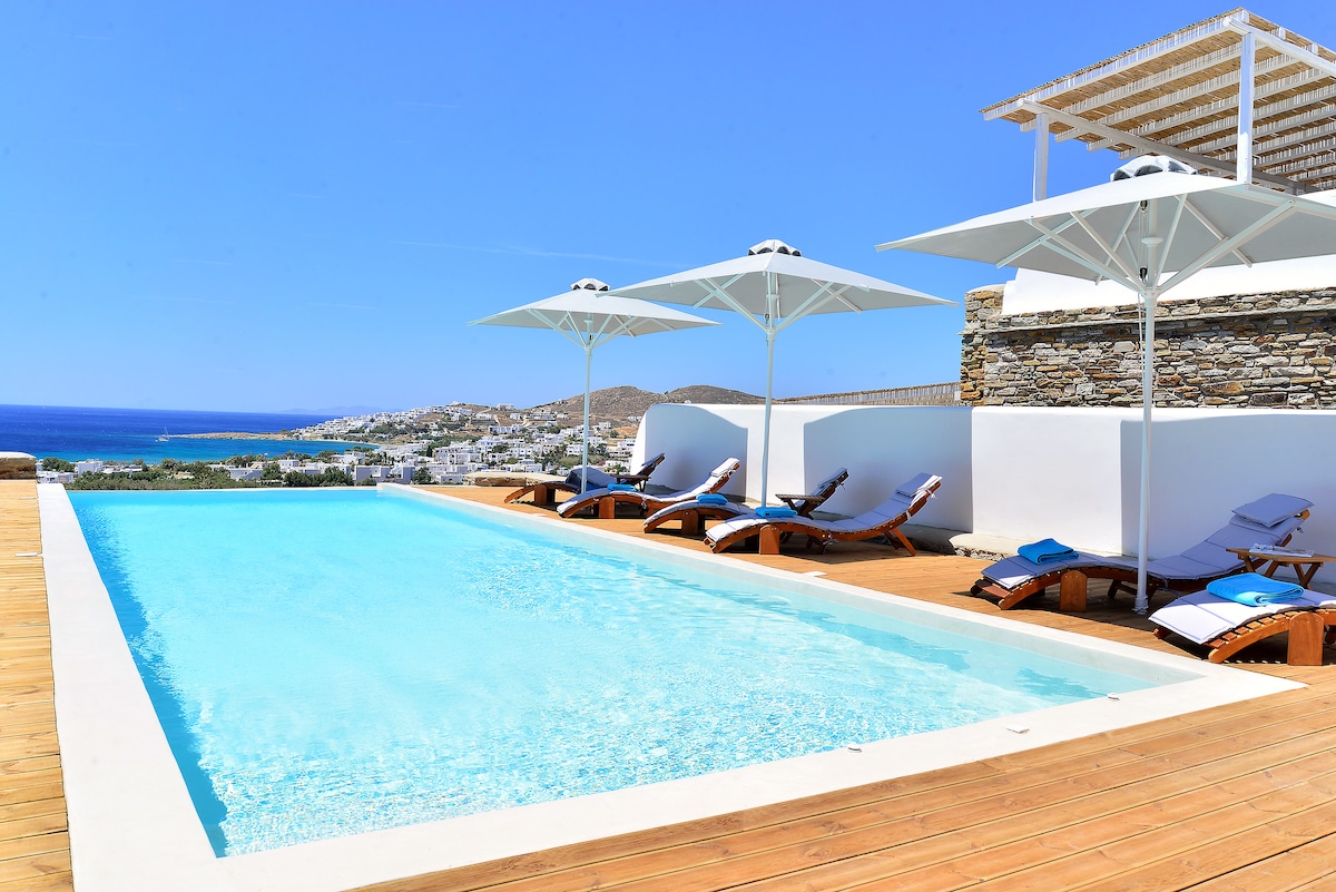 A sunlit outdoor pool surrounded by wooden decking features several loungers with umbrellas for shade. Views of the sea and Tinos island's landscape are visible in the background, enhancing the serene setting.