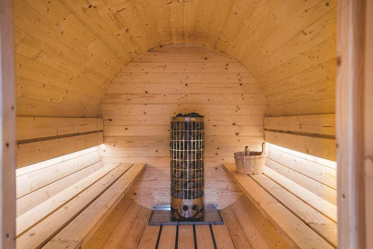 Inside the sauna, warm wooden walls and benches create a cozy atmosphere. A cylindrical stove sits centrally, surrounded by slatted seating on either side. Soft lighting enhances the tranquil environment, inviting relaxation.