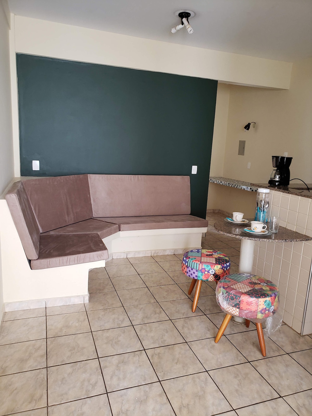 A cozy dining area features a curved, upholstered bench integrated into the wall, complemented by two colorful stools with patchwork designs. A small countertop with a coffee maker and two cups sits nearby, against a backdrop of neutral-toned tiles and a dark accent wall.