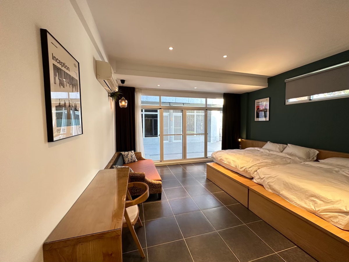 A spacious room features a large bed with white bedding, accented by a wooden platform. A comfortable seating area is present with decorative cushions. Natural light floods through floor-to-ceiling glass doors, leading to a private outdoor space, while artwork decorates the walls.