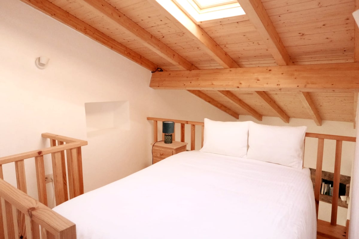 A cozy sleeping area is highlighted by a spacious bed adorned with crisp white linens. Natural wood beams run across the ceiling. A bedside table is positioned near a small lamp, and a skylight above allows natural light to illuminate the room.