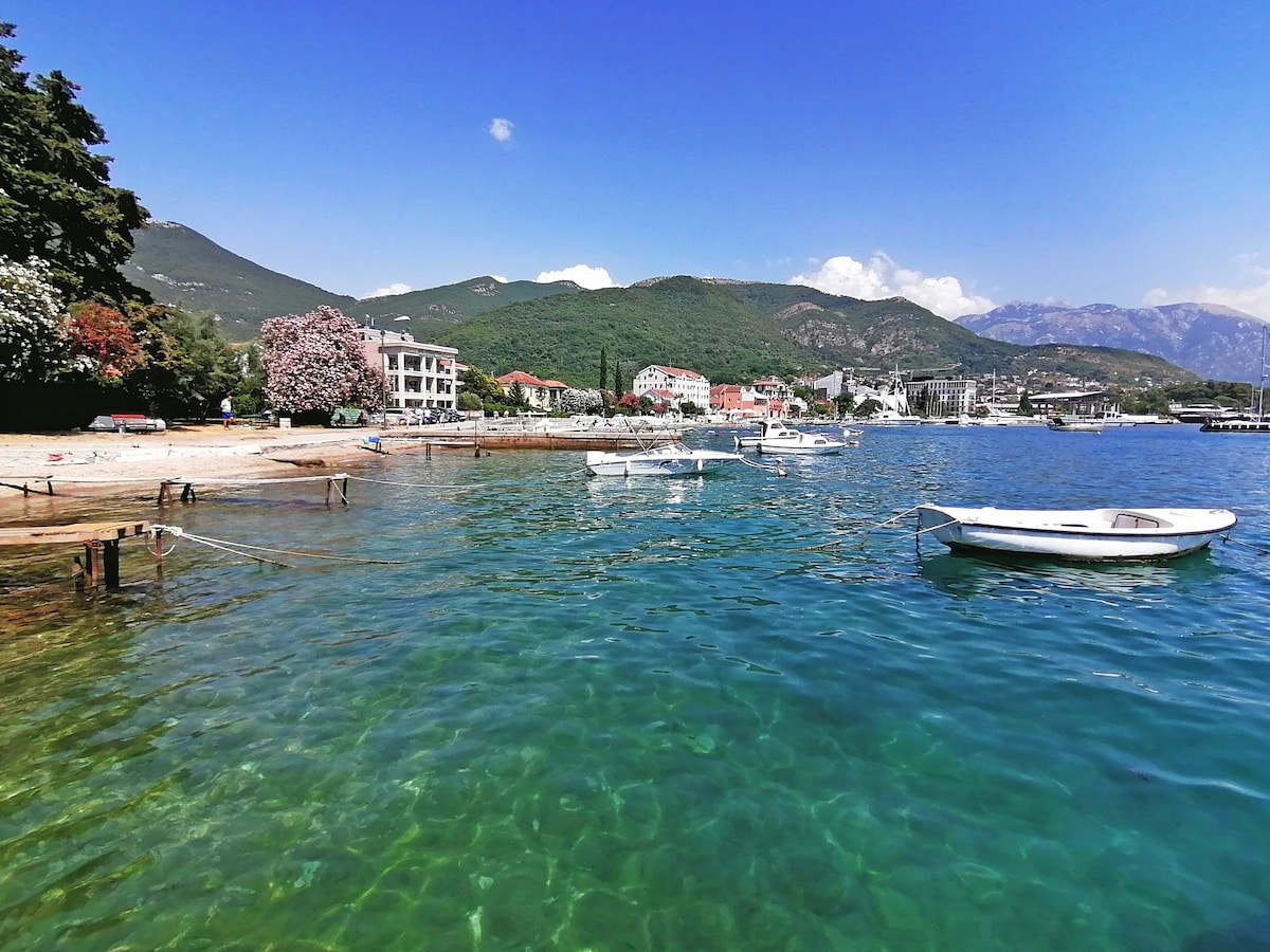 A serene waterscape is presented, with crystal-clear water reflecting the vibrant blue sky. Small boats are anchored along the shore, and the coastline features a mix of sandy and rocky areas surrounded by lush greenery and distant mountains.