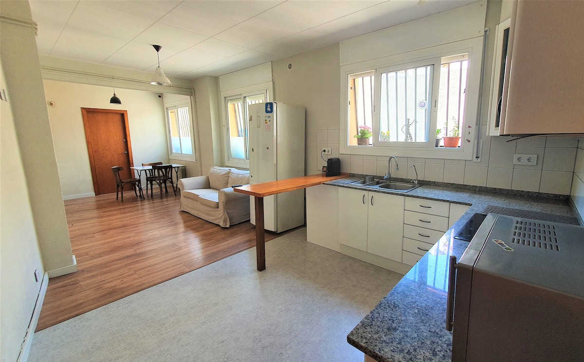 A modern kitchen area features a sleek countertop and ample cabinetry. A table with four chairs is visible in the background, alongside a cozy seating area with a light-colored couch. Large windows provide natural light, enhancing the spacious layout.