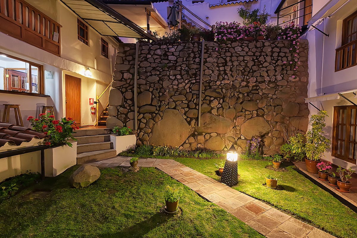 A well-kept garden is surrounded by stone walls and features a pathway leading to various potted plants. Soft lighting illuminates the area, enhancing the natural beauty of the lush greenery and colorful flowers.